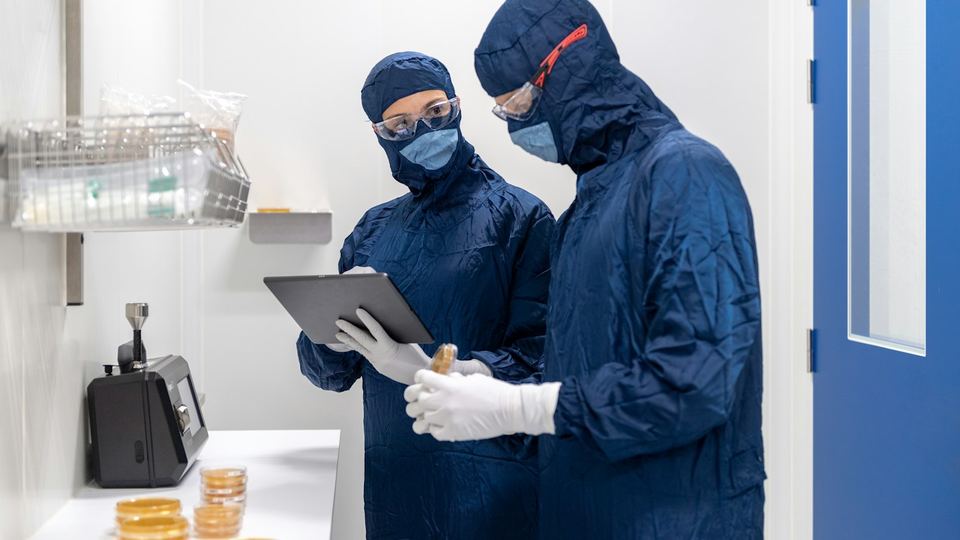 Two lab scientists in cleanroom suits reviewing results on a tablet